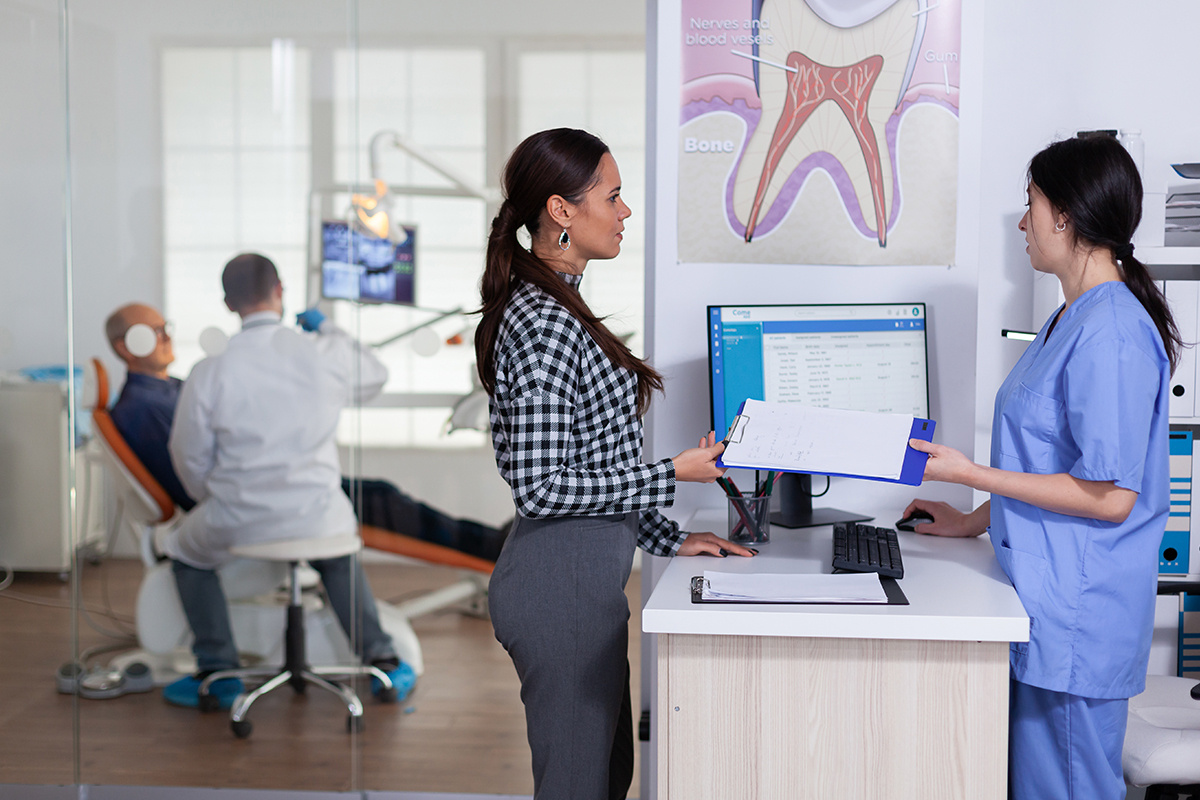 A patient talking to a receptionist while a dentist is seeing another patient in the background.