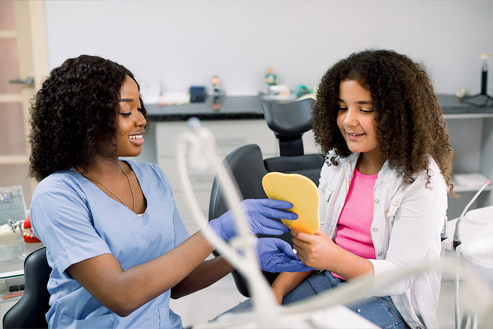 Teen girl, looking at the mirror with toothy smile, sitting on the dental chair while smiling dentist showing the result of treatment